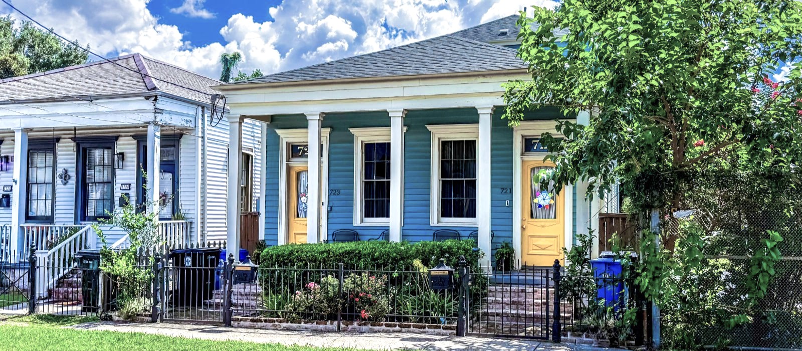 A row of New Orleans houses on a sunny day — blue siding, yellow doors, ornamental ironwork along the sidewalk.
